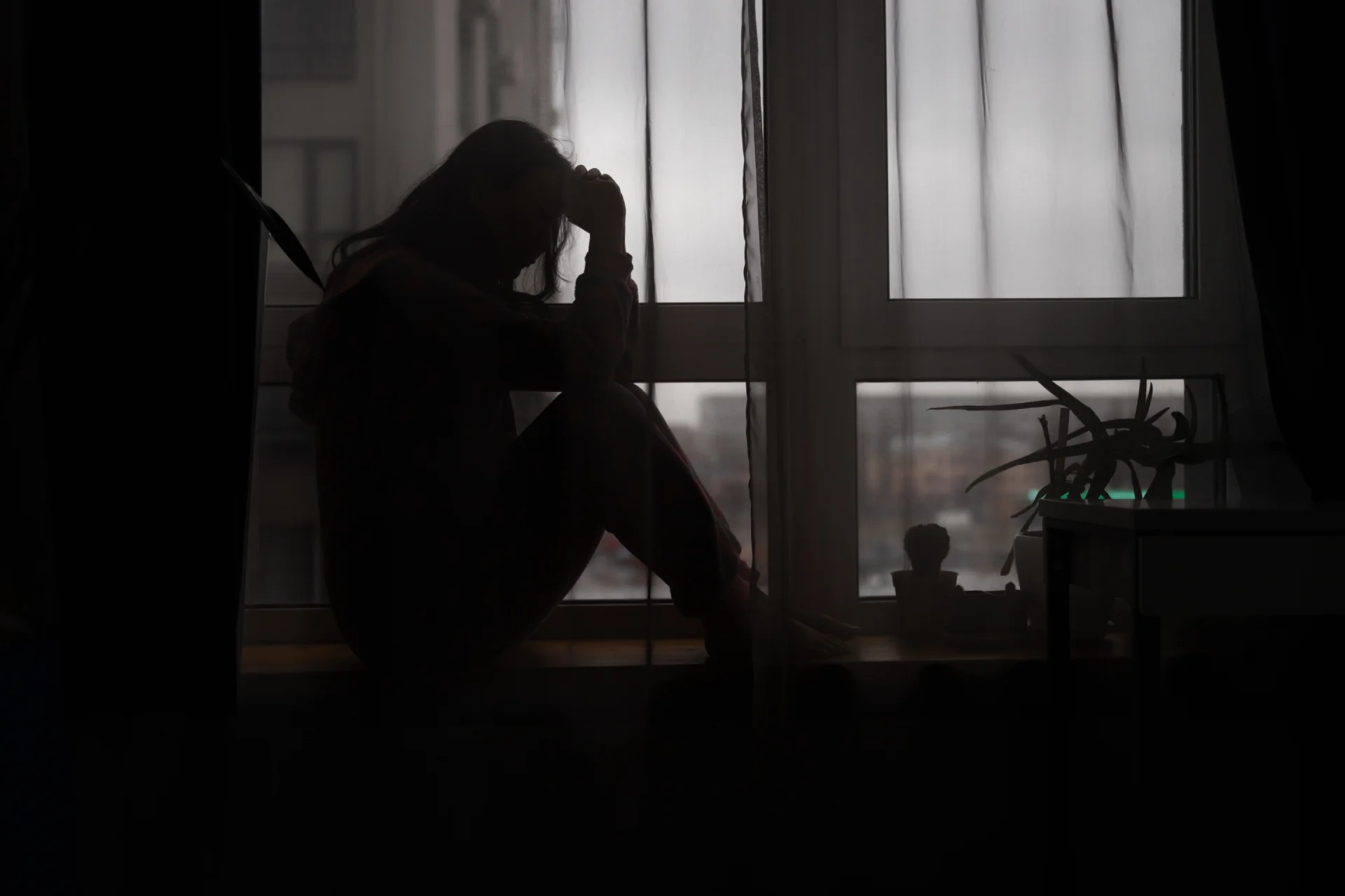 Woman sitting by the window, dark room, having negative thoughts, feeling sad and depressed.